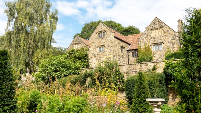 Late summer, blue sky with white clouds. The stone exterior and red tiled roof of Washington Old Hall viewed from gardens below with plants in bloom, topiary shrubs and a stone sundial in the foreground.